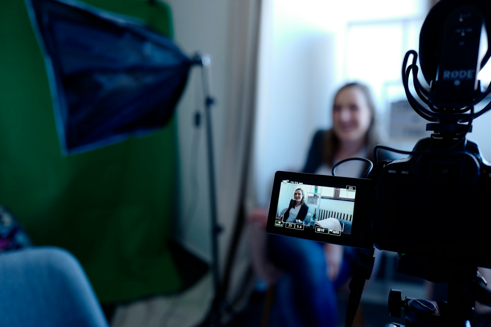 A camera with a microphone recording a smiling presenter in a small studio with a green screen